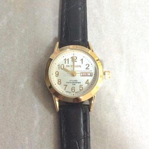 MIYKON Damen-Golduhr mit rundem, weißem Day & Datum-Zifferblatt mit nummerierten Goldstunden auf einem schwarzen Lederband.