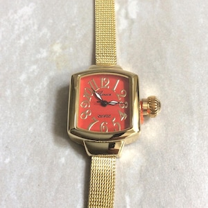 May include: Gold-toned quartz watch with a square face and a mesh band. The watch face is a vibrant orange with gold numerals and hands. The watch has a crown on the side and the word "Quartz" is printed on the face.