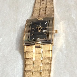 May include: Gold-toned wristwatch with a rectangular black face. The watch features a gold band with a textured, brick-like pattern. The watch face displays the brand name "SUNDOMI" and the words "WATER RESISTANT".