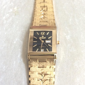 Puede incluir: Reloj de pulsera dorado con esfera negra rectangular. La correa y la caja son doradas, con un diseño texturizado. La esfera muestra la marca "Sunborn", el día y la fecha. La correa mide aproximadamente 2 cm de ancho.