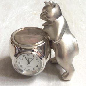 May include: A silver-toned metal desk clock featuring a round face with black numerals and the text Geneva Quartz. The clock is integrated into a rounded base next to a standing silver-toned cat figurine that rests its front paws on the clock top.