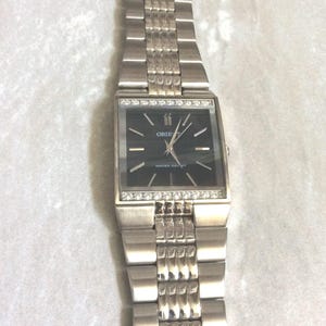 Puede incluir: Reloj de pulsera plateado con una esfera negra rectangular. El reloj presenta una correa plateada y un bisel adornado con pequeñas piedras transparentes. La esfera del reloj muestra la marca "ORIENT" y las palabras "WATER RESIST".