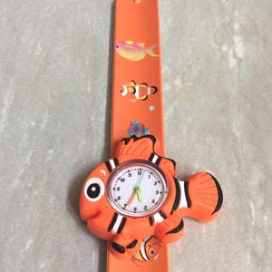May include: Orange children's watch with a clownfish design. The watch face is round with white numbers and hands. The band is orange with fish illustrations.