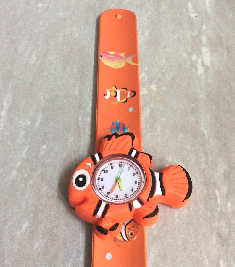 May include: Orange children's watch with a clownfish design. The watch face is round with white numbers and hands. The band is orange with fish illustrations.