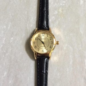 May include: A gold-coloured wristwatch with a black leather strap. The watch face displays the brand name "MIYKON" and features black numerals on a gold background. The strap has a textured, crocodile-like pattern.