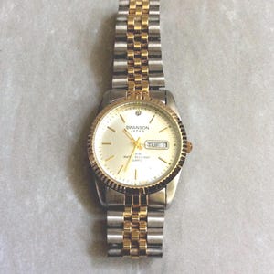 May include: A Swanson Japan quartz watch with a silver and gold-tone band and bezel. The watch face is cream-colored with gold hands and markers, and displays the day and date. The watch is water-resistant.