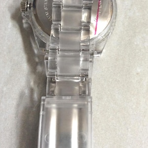 Puede incluir: Un reloj de pulsera transparente y claro con una correa a juego. La esfera del reloj es plateada con manecillas y marcadores negros. La correa est&aacute; hecha de eslabones de pl&aacute;stico transparente. El reloj se ve desde atr&aacute;s.