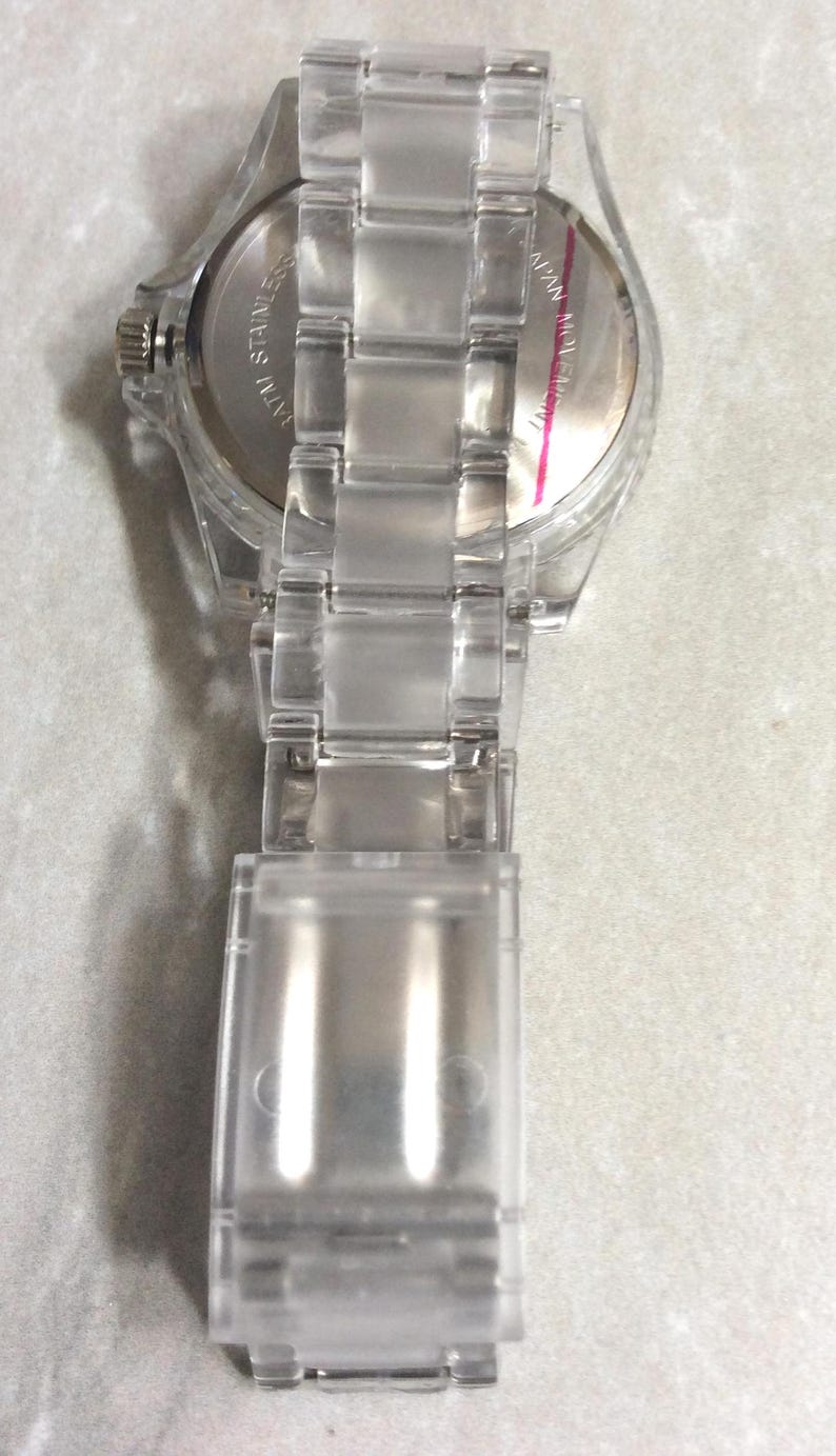 Puede incluir: Un reloj de pulsera transparente y claro con una correa a juego. La esfera del reloj es plateada con manecillas y marcadores negros. La correa est&aacute; hecha de eslabones de pl&aacute;stico transparente. El reloj se ve desde atr&aacute;s.