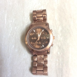 May include: A rose gold-toned wristwatch with a matching metal band. The watch face has a dark brown background with gold-colored hands and markers. The watch features multiple sub-dials and a date display.