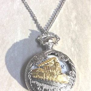 May include: Silver pocket watch pendant with a gold-coloured train design. The watch has a white clock face with black numerals and hands. The pendant hangs from a silver chain, with a vintage or antique aesthetic.