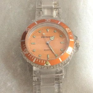 May include: A clear plastic watch with an orange face and bezel. The watch face displays the word "Vensal" and "Quartz". The watch has a clear plastic band. The bezel has the numbers 20, 30, and 40.