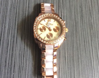 May include: A rose gold wristwatch with a cream-colored face and a bezel encrusted with small, clear crystals. The watch has a matching rose gold and white band. The watch face has three smaller dials.
