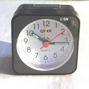 May include: A black square quartz alarm clock with a white face. The clock face has black numerals, a red second hand, and green and yellow hands. The clock reads "QUARTZ" and has the text "L-SN" on the top right.