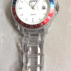 Puede incluir: Reloj de cuarzo transparente con esfera blanca, con la marca "ASPEN". El reloj tiene un bisel rojo y azul y una correa transparente. La esfera muestra la hora y la palabra "QUARTZ".