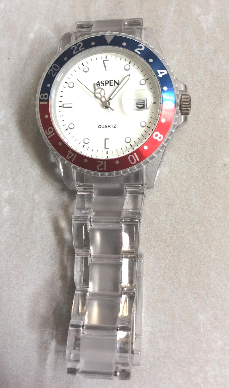 Puede incluir: Reloj de cuarzo transparente con esfera blanca, con la marca "ASPEN". El reloj tiene un bisel rojo y azul y una correa transparente. La esfera muestra la hora y la palabra "QUARTZ".