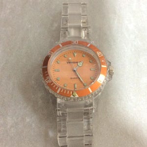 Puede incluir: Un reloj de plástico transparente con esfera y bisel naranjas. La esfera del reloj tiene las palabras "Versai" y "Quartz" impresas. La correa del reloj también es de plástico transparente.