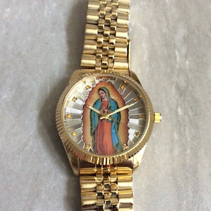 May include: Gold-toned wristwatch featuring a detailed image of the Virgin Mary on the watch face. The watch has a gold band and a textured bezel. The watch face is surrounded by gold accents.