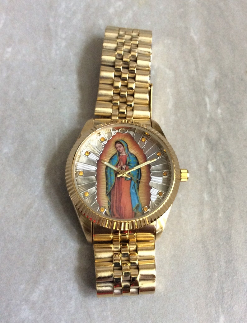 May include: Gold-toned wristwatch featuring a detailed image of the Virgin Mary on the watch face. The watch has a gold band and a textured bezel. The watch face is surrounded by gold accents.