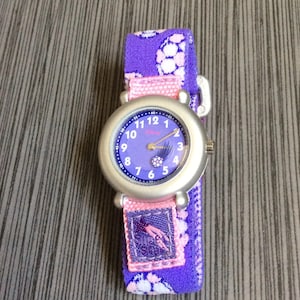 Pode incluir: Um relógio de pulso prateado com mostrador roxo e pulseira a condizer. O mostrador do relógio tem algarismos brancos e um gráfico de bola de futebol. A pulseira é roxa com um padrão floral branco e uma correia rosa.