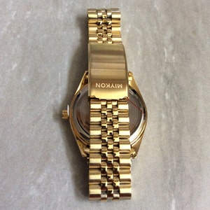 May include: A gold-toned wristwatch with a matching band. The watch face is visible, and the band is partially open. The clasp is engraved with the word "MIYKON". The watch has a classic, elegant design.