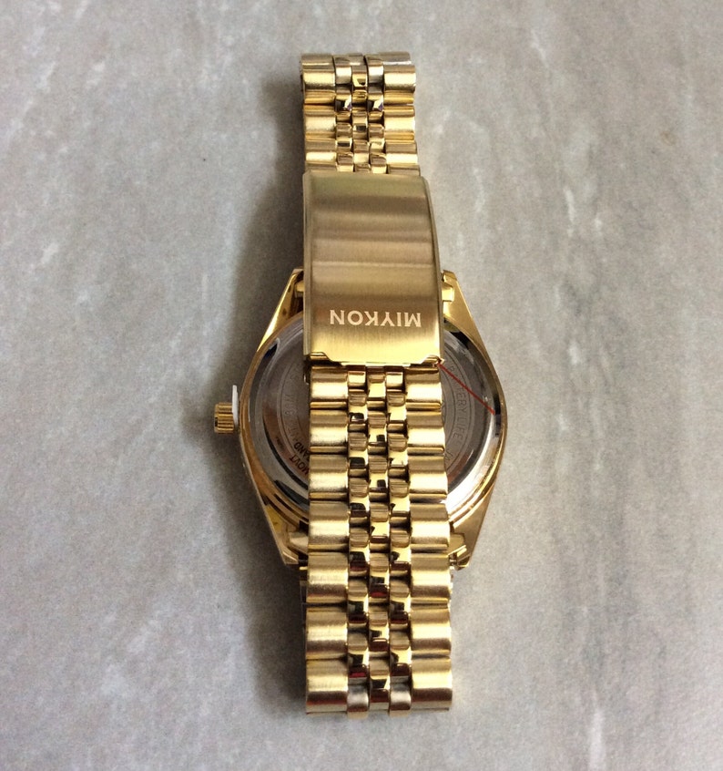 May include: A gold-toned wristwatch with a matching band. The watch face is visible, and the band is partially open. The clasp is engraved with the word "MIYKON". The watch has a classic, elegant design.