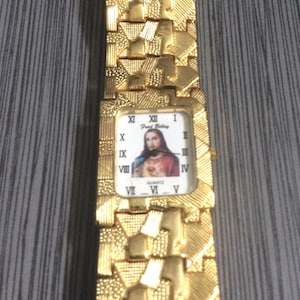 May include: Gold-coloured wristwatch with a textured strap and a rectangular face. The watch face displays a portrait of Jesus Christ and Roman numeral hour markers. The watch is quartz.