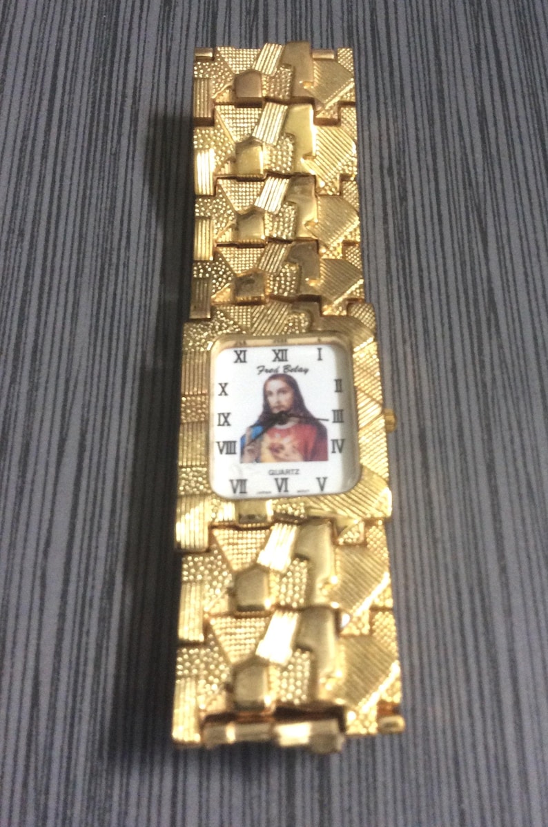 May include: Gold-coloured wristwatch with a textured strap and a rectangular face. The watch face displays a portrait of Jesus Christ and Roman numeral hour markers. The watch is quartz.