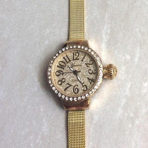 Puede incluir: Un reloj de pulsera dorado con una correa de malla. La esfera del reloj está rodeada por un anillo de cristales transparentes y presenta grandes números negros. La esfera del reloj tiene un aspecto brillante y texturizado. El reloj tiene una pequeña corona texturizada.