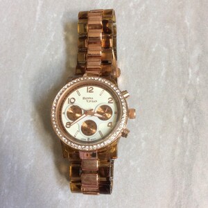 May include: A rose gold wristwatch with a cream-colored face and a bezel encrusted with small, clear crystals. The watch features three sub-dials and the text "Donna Vivian". The band is a matching rose gold color.