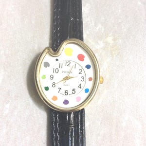 May include: A gold-toned wristwatch with a white face featuring colorful paint splotches and black numerals. The watch has a black leather band and the word "FINESSE" is printed on the watch face.