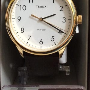 Könnte beinhalten: Eine goldfarbene Timex-Uhr mit weißem Zifferblatt, schwarzen Zeigern und einem braunen Lederarmband. Das Zifferblatt zeigt das Timex-Logo und das Wort "INDIGLO". Die Uhr wird in einer roten Box mit dem Timex-Logo präsentiert.
