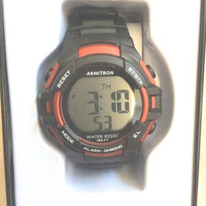 May include: Black and red Armitron Pro Sport digital watch. The watch face displays the time 3:10:53. Features include water resistance to 165 feet, alarm, and chronograph functions. The watch is in its original packaging.