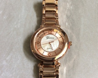 May include: Rose gold Geneva quartz watch with a white mother-of-pearl face. The watch has a round face with gold-tone hands and hour markers. The watch band is made of rose gold links.