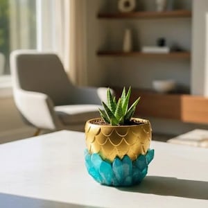 May include: A decorative succulent plant in a unique pot. The pot has a gold-colored, scale-like upper section and a turquoise, crystal-like base. The plant has green, pointed leaves and is set on a white table.