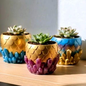 May include: Three decorative succulent planters. The planters are gold with a scale-like texture and a base of blue, purple, and gold crystal-like formations. Each planter holds a green succulent plant. The planters are on a wooden surface.