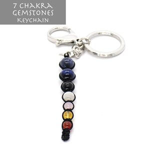 May include: A silver keychain with a loop and a chain attached to a string of seven chakra gemstones. The gemstones are blue, black, white, pink, yellow, orange, and red.