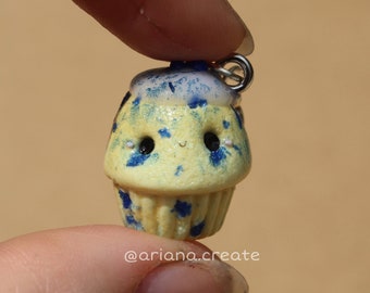 May include: A small yellow cupcake with blue frosting and sprinkles. The cupcake has a cute cartoon face with black eyes and a smile. The cupcake is attached to a silver chain.