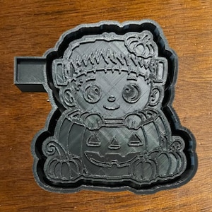 May include: Black plastic cookie cutter in the shape of a cartoon Frankenstein's monster holding a jack-o'-lantern. The cutter has a handle on the side and is designed for making Halloween-themed cookies. The design includes details like stitches and pumpkin features.