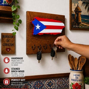May include: A rectangular stained wood wall-mounted key holder featuring a cutout of the Puerto Rican flag. The wooden board includes five black metal hooks for hanging keys. The flag design displays a blue triangle with a white star and red and white stripes.