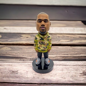 May include: A bobblehead figurine of a person with a brown complexion wearing a green, brown, and black camouflage hoodie and grey and orange sneakers. The figurine is standing on a black base.