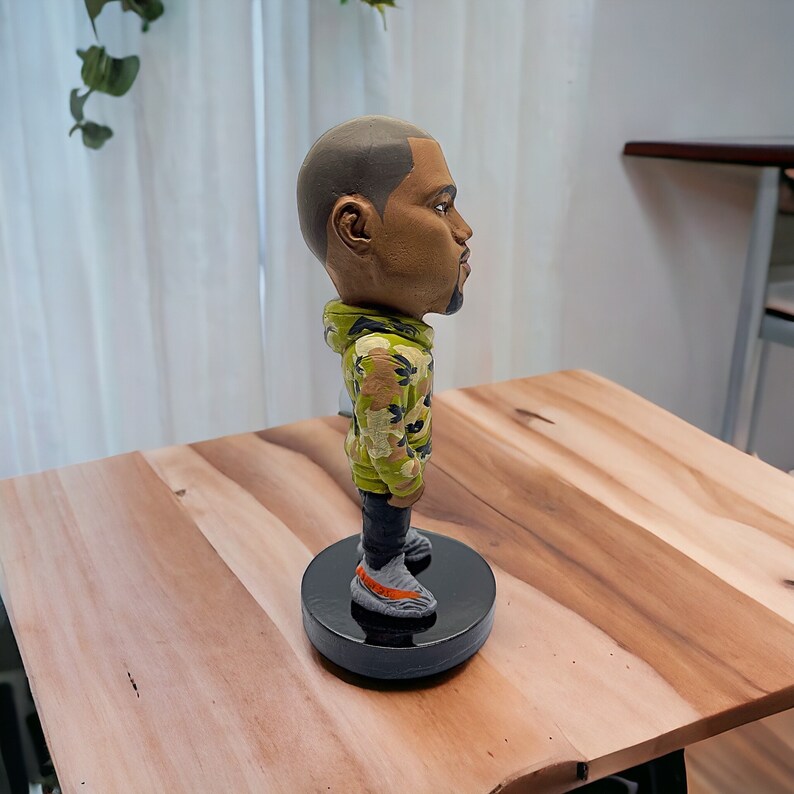 May include: A figurine of a person wearing a green, brown, and black camouflage jacket and grey shoes. The figurine is standing on a black circular base.