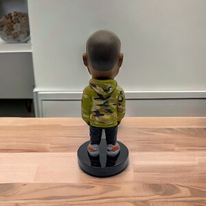 May include: A figurine of a person wearing a green, black, and brown camouflage hoodie and black pants. The figurine is standing on a black base.
