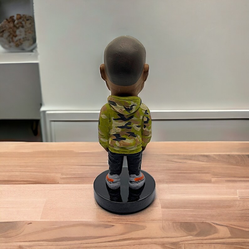 May include: A figurine of a person wearing a green, black, and brown camouflage hoodie and black pants. The figurine is standing on a black base.