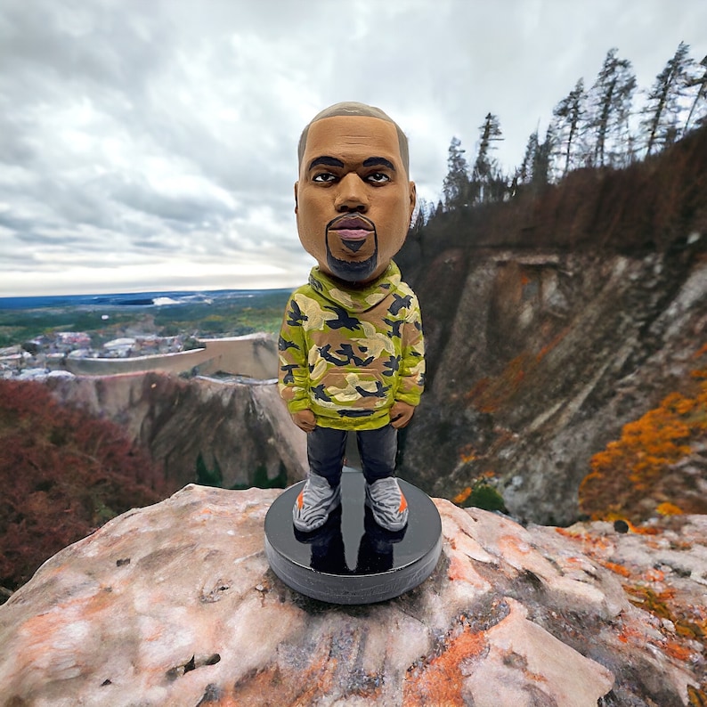 May include: A bobblehead figurine of a person wearing a camouflage hoodie and black pants. The figurine is standing on a black base and is positioned on a rocky surface with a view of a valley in the background.