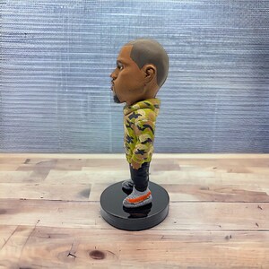 May include: A small figurine of a person wearing a camouflage jacket and grey sneakers. The figurine is standing on a black base.