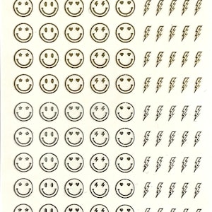 以下が含まれることがあります： スマイリーフェイス、ハート、稲妻、星形のゴールドのネイルステッカー。ステッカーは白い背景に貼られています。www.liquidglamnailsupply.com