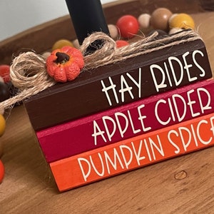 May include: Stacked wooden blocks with fall-themed text: "Hay Rides", "Apple Cider", and "Pumpkin Spice". The blocks are painted in shades of brown, red, and orange. A small orange felt pumpkin is attached to the top block with twine.