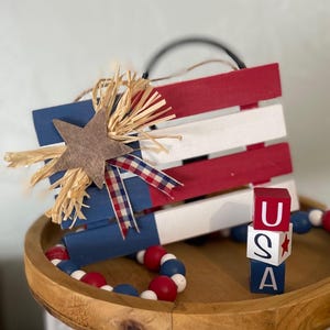 May include: A wooden sign painted in red, white, and blue with a star and a bow. The sign is sitting on a wooden tray with red, white, and blue beads. There are three wooden blocks stacked on top of each other that spell "USA".
