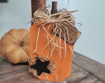 Può includere: Una sagoma di zucca in legno dipinta di arancione con un gambo a ramoscello marrone scuro, un fiocco in rafia beige e una stella nera. Un piccolo cartellino rettangolare in carta con bordi bruciati riporta la scritta Happy Fall in inchiostro nero.