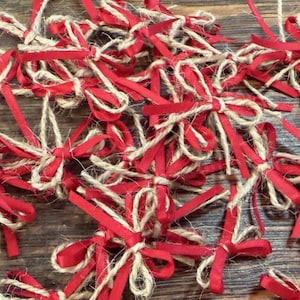 May include: A collection of small decorative bows. Each bow features a red satin ribbon tied with natural twine. The bows are scattered on a wooden surface, creating a festive and handcrafted appearance.